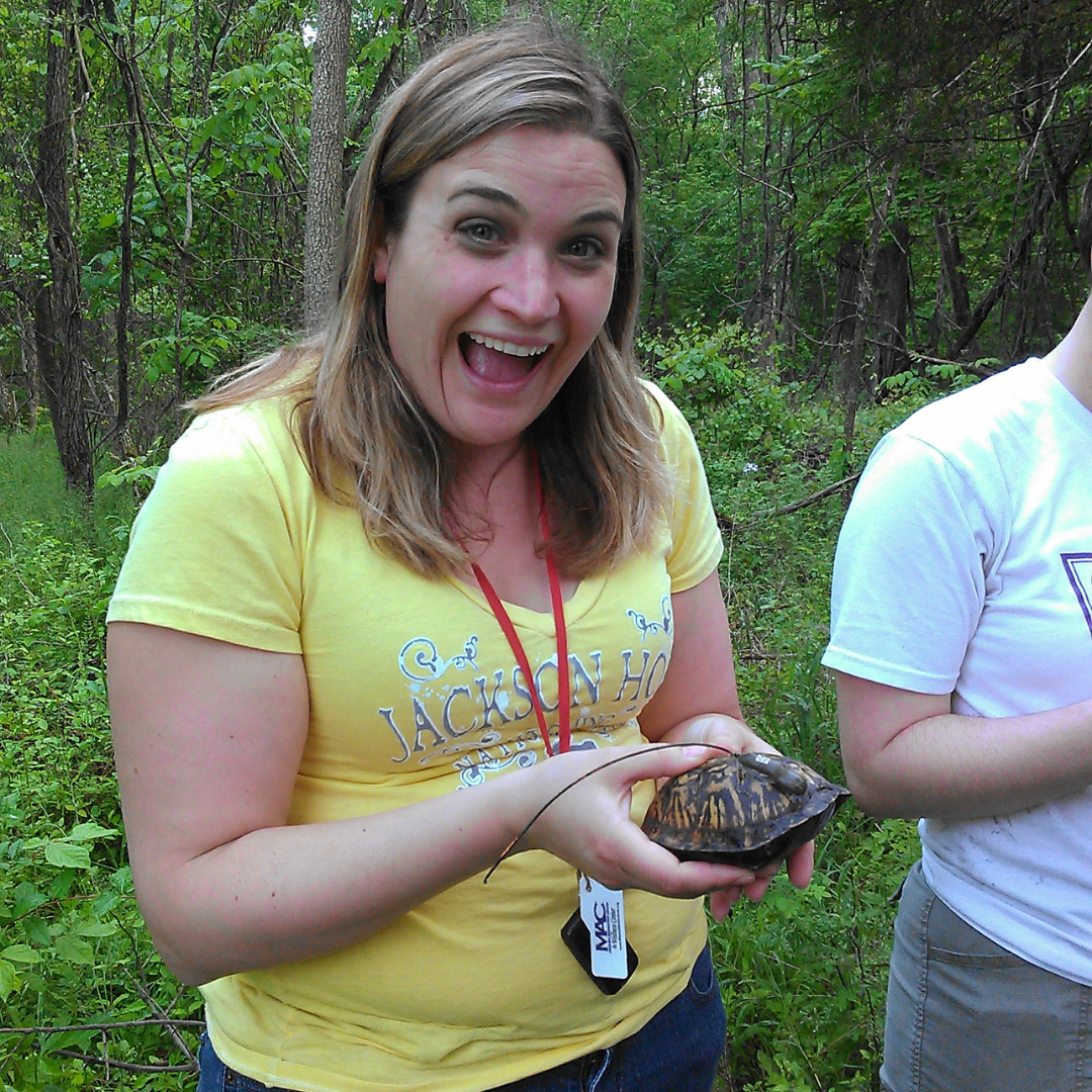 A white woman in a yellow tshirt grins widely as she holds a box turtle